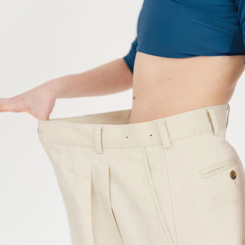 service-1-1 Unrecognizable slim female with bare belly wearing white oversized pants after weight loss on white background in modern light studio